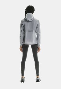 Veste grise imperméable avec capuche, manches longues et poche arrière, associée à des leggings noirs et des chaussures de sport grises.