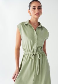 Svijetlozelena, bezrukavna haljina na dugme s ovratnikom, struk koja se može zategnuti i prozračna tekstura tkanine. Glatka završna obrada, ležeran dizajn.
