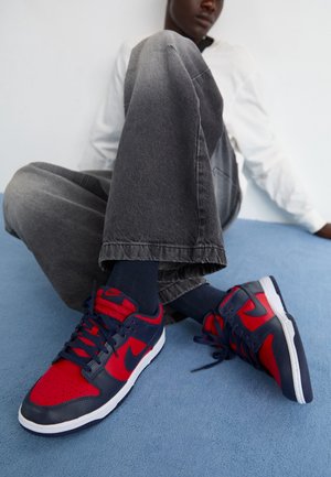 Personne portant un jean gris et une chemise blanche assise sur un sol bleu avec des baskets rouges et bleu marine visibles.