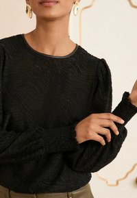 Maglione nero con texture a onde, colletto rotondo e maniche arricciate con polsini a volant. Modello in posa con le mani parzialmente visibili.