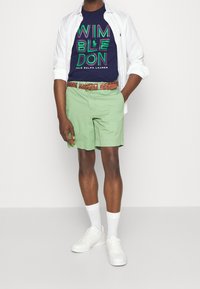 Gröna shorts, marinblå T-shirt med "WIMBLEDON"-grafik, vit skjorta med knappar, flätat brunt bälte, vita sneakers och vita strumpor.