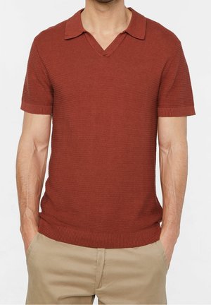 Mężczyzna w rdzawej, krótkorękawowej, dzianinowej koszulce polo i beżowych spodniach, z rękami w kieszeniach, na tle jednolitego jasnego tła.