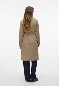 Trench coat beige con maniche lunghe e vita con cintura, caratterizzato da una texture liscia e un taglio classico. Indossato sopra jeans blu scuro.