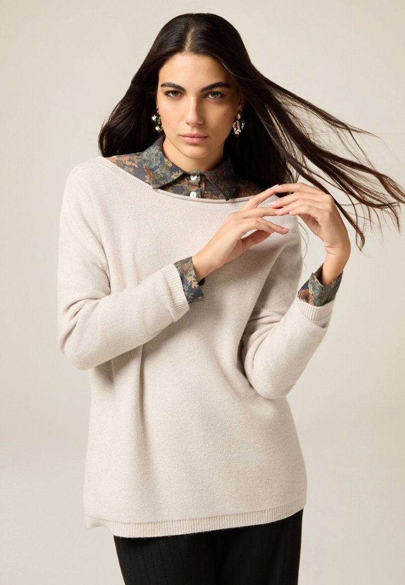 Helles Beigesweatshirt mit weitem Ausschnitt und weicher Textur, über einem dunklen, floral gemusterten Hemd mit Kragen getragen, kombiniert mit schwarzen Nadelstreifen-Hosen.