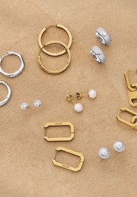 Orecchini in oro e argento di vari design, tra cui cerchi, perni e accentuazioni testurizzate, disposti su una superficie beige.