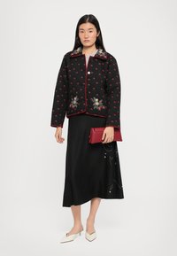 Mustat tikatut takki, jossa on punaisia kukka-ompeluja, nappisuljenta ja kontrastinen punainen tikkaus. Päällä mustan hameen ja punaisen clutch-laukun kanssa.