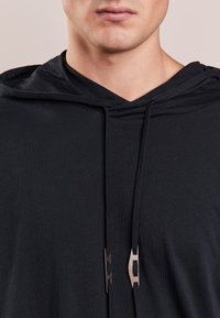 Musta vetoketjullinen huppari, jossa V-pääntie, valmistettu pehmeästä materiaalista, mustat kiristysnauhat ja metalliset yksityiskohdat nauhojen päissä.