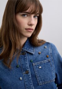Denimjakke i blå med lys vask, med uregelmæssigt placerede farverige sten på brystet og kraven samt en lomme med knap.