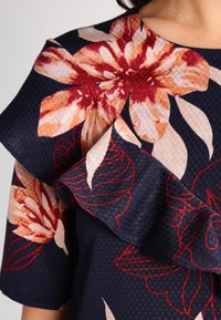 Top a fantasia floreale con grandi fiori arancioni e crema su uno sfondo blu scuro, dotato di un colletto e maniche arricciati e testurizzati.