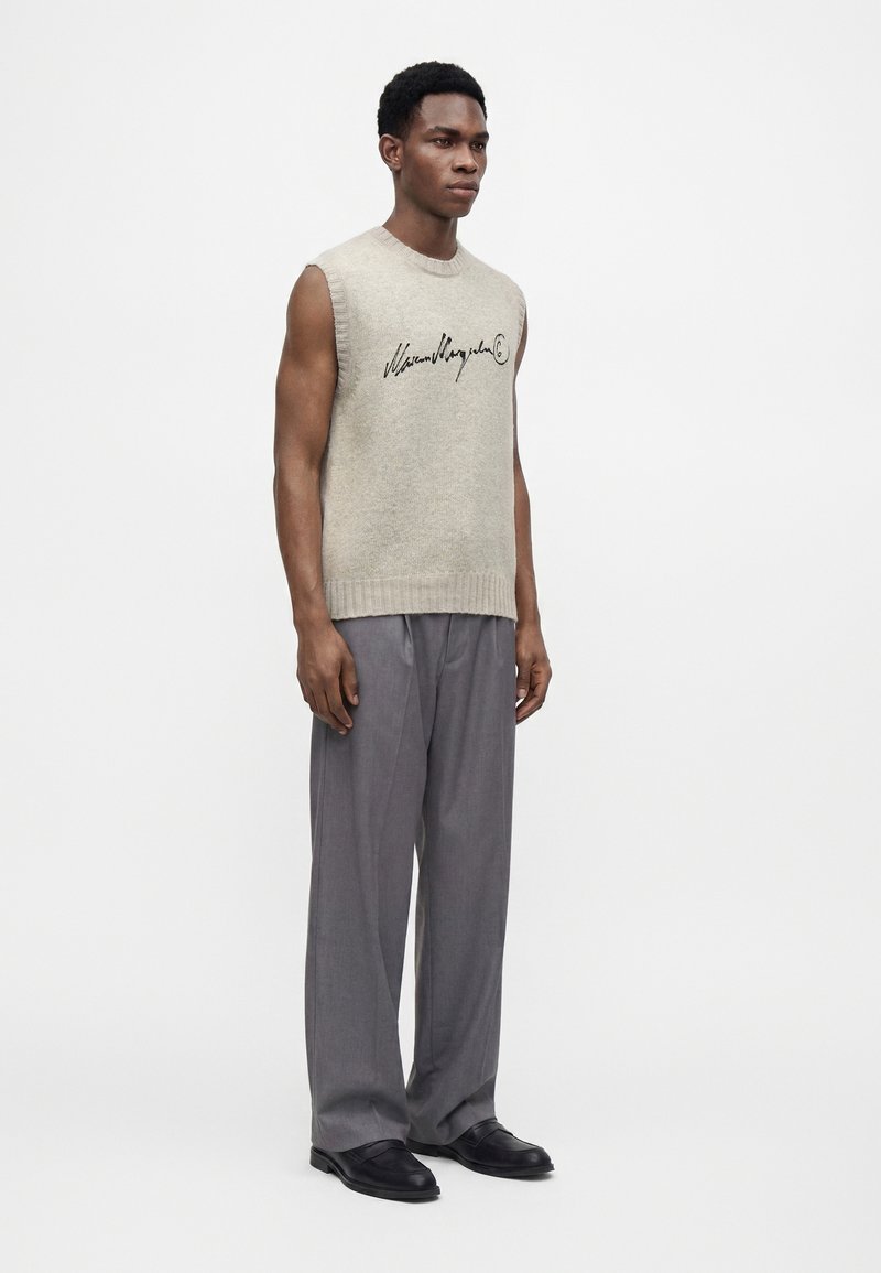 Junger Mann steht, trägt einen ärmellosen beigen Pullover mit schwarzer Schrift und graue weite Hosen mit schwarzen Schuhen.