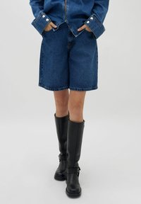 Denim kratke hlače sproščenega kroja, srednje dolžine do stegen, svetlo modre pranja, sprednje žepke, v kombinaciji z črnimi usnjenimi škornji do kolen.