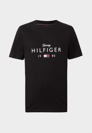 T-shirt preta de manga curta com texto branco "Tommy Hilfiger 1985" e logotipo da bandeira em vermelho, branco e azul centrado na parte da frente.