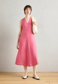 Roza haljina od lana s V-izrezom, bez rukava, A-linija silueta i dužina do sredine lista. Model drži kremastu torbu.