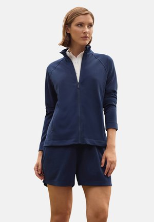 Mujer con chaqueta azul marino con cremallera sobre una camiseta blanca y pantalones cortos a juego en azul marino, de pie con los brazos relajados contra un fondo claro y liso.