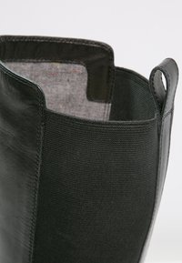 Primer plano de la parte superior de una bota de cuero negro que muestra el panel lateral elástico y la lengüeta contra un fondo blanco.