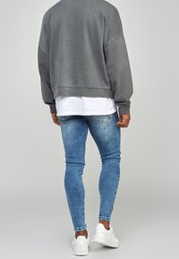Sudadera gris de gran tamaño con dobladillo y puños acanalados, superpuesta sobre una camisa blanca, combinada con jeans azules ajustados y zapatillas blancas.
