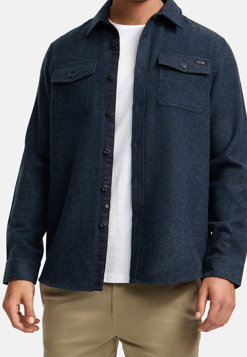 Homme portant une veste-chemise en laine bleu foncé déboutonnée par-dessus un t-shirt blanc et un pantalon beige, mains relâchées le long du corps.
