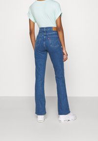 Jeans de mezclilla azul de talle alto con pierna acampanada, que cuentan con un parche de cuero marrón en la parte trasera y una etiqueta de logo roja en el bolsillo derecho.
