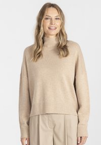 Beżowy sweter z golfem, z ribbanym wykończeniem u dołu i na mankietach, o luźnym kroju i miękkiej fakturze. W zestawie z jasnobeżowymi spodniami.