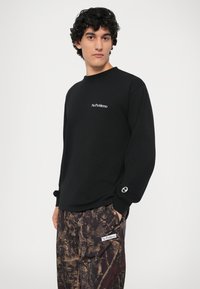 Zwarte sweatshirt met lange mouwen met de tekst "NoProblemo" in het wit op de borst. Beschikt over een manchet met een cirkelvormig logo-detail.