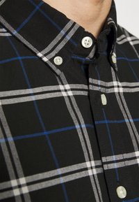 Zwart en geruit shirt met witte en blauwe ruitpatronen, knoop-om-kraag en lichtgekleurde knopen op een textuele stof.