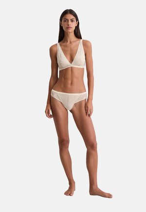 Frau, die barfuß steht und ein beiges Spitzen-Bralette sowie passende hochgeschnittene Slips vor einem schlichten weißen Hintergrund trägt.