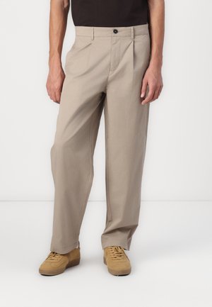 Pantalon beige coupe large avec des plis, porté avec des baskets couleur cuir clair et un haut noir, visible de la taille jusqu'aux pieds.