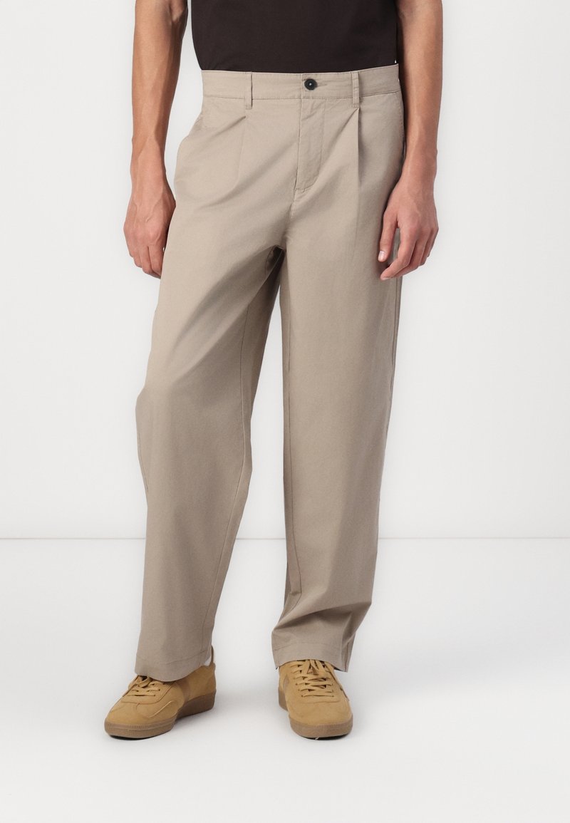 Pantalon beige coupe large avec des plis, porté avec des baskets couleur cuir clair et un haut noir, visible de la taille jusqu'aux pieds.
