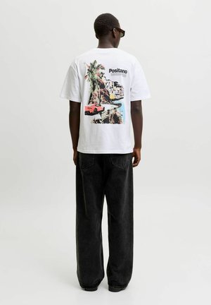 Personne portant un t-shirt blanc avec un imprimé coloré de la côte de Positano et un jean noir à jambes larges, debout devant un fond uni.