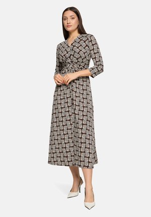Kvinde iført en sort og hvid midi slå-om kjole med geometrisk mønster og 3/4 ærmer samt hvide spidse højhælede sko, stående mod en ensfarvet baggrund.