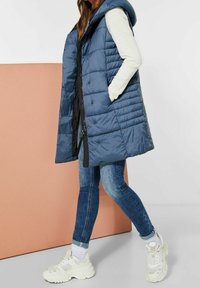 Blå vadderad väst med huva, med dragkedja framtill, stora sidofickor och quiltat mönster, kombinerad med mörka jeans och vita sneakers.