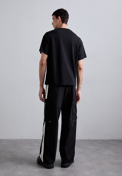Man standing against plain wall, wearing black T-shirt and black cargo pants with white side stripes, viewed from the back.