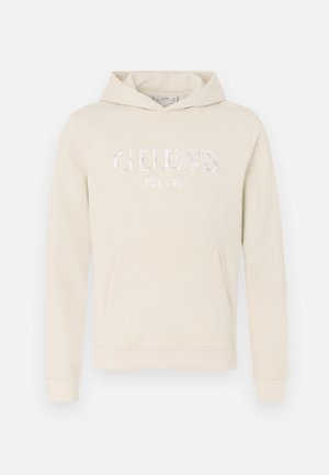 Beige hættetrøje med en frontlomme, der har et broderet "GUESS EST.1981" logo i hvid. Blød tekstur og justerbar hætte. Lange ærmer.