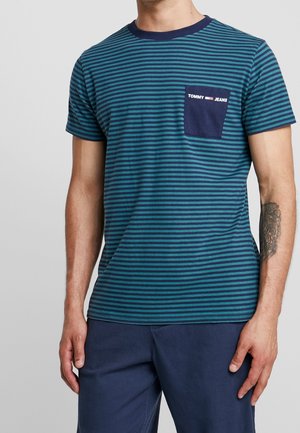 Mężczyzna ubrany w krótki rękaw w paski w kolorze mięty i granatu, z granatową kieszenią z napisem "Tommy Jeans" oraz granatowe spodnie.