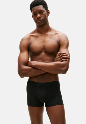 Hombre musculoso con los brazos cruzados, vestido con bóxers negros, de pie frente a un fondo blanco liso.