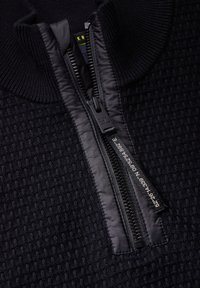 Svart tröja med en strukturerad stickad mönster och en slät svart zipkrage. Inkluderar en dragkedjepull med koordinater tryckta längs dragkedjan.