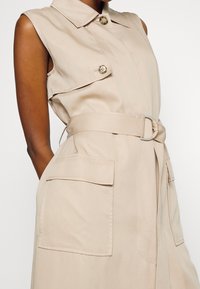Beżowa sukienka bez rękawów z kołnierzem, z dwoma przednimi kieszeniami i dopasowanym paskiem, wykonana z gładkiej tkaniny z subtelnymi detalami przeszyć.