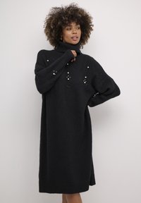 Melna adīta kleita ar augstu kakla izgriezumu, dekorēta ar pērlītēm un brīvu siluetu.