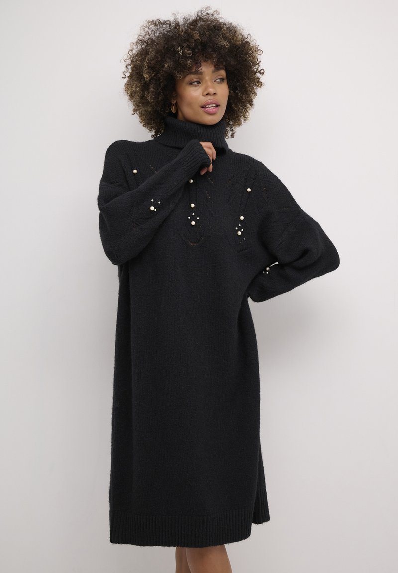 Melna adīta kleita ar augstu kakla izgriezumu, dekorēta ar pērlītēm un brīvu siluetu.