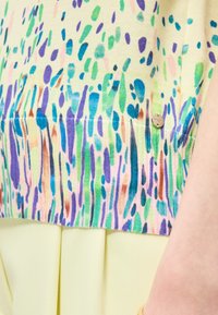 Κοντινή εικόνα πολύχρωμου υφάσματος με αφηρημένες κηλίδες και ρίγες, συνοδευόμενο από ανοιχτό κίτρινο πλισέ κάτω μέρος και ορατό χέρι.