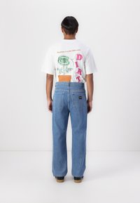 Hvid T-shirt med grafisk print på ryggen og blå denim bukser med vide ben. Bemærkelsesværdige detaljer inkluderer syet lap og branding på bukserne.
