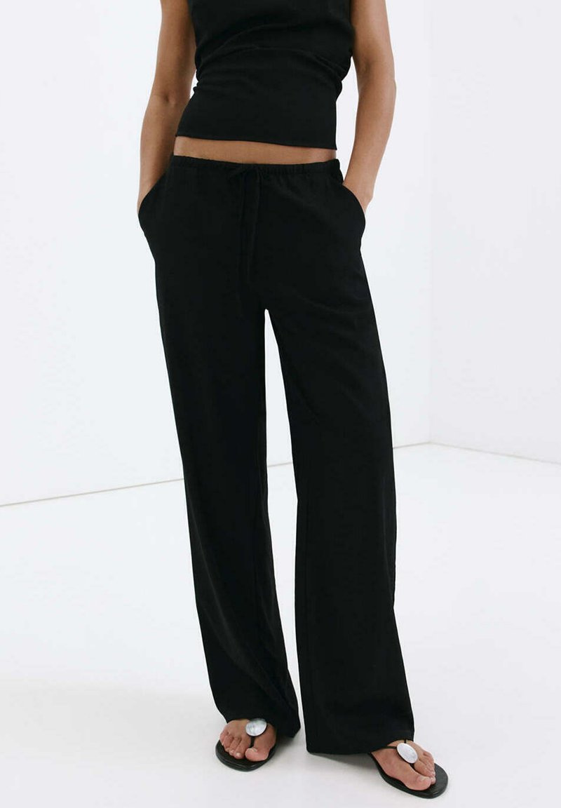 Personne portant un pantalon noir à jambes larges et un haut noir sans manches, les mains dans les poches, debout pieds nus sur un sol blanc.