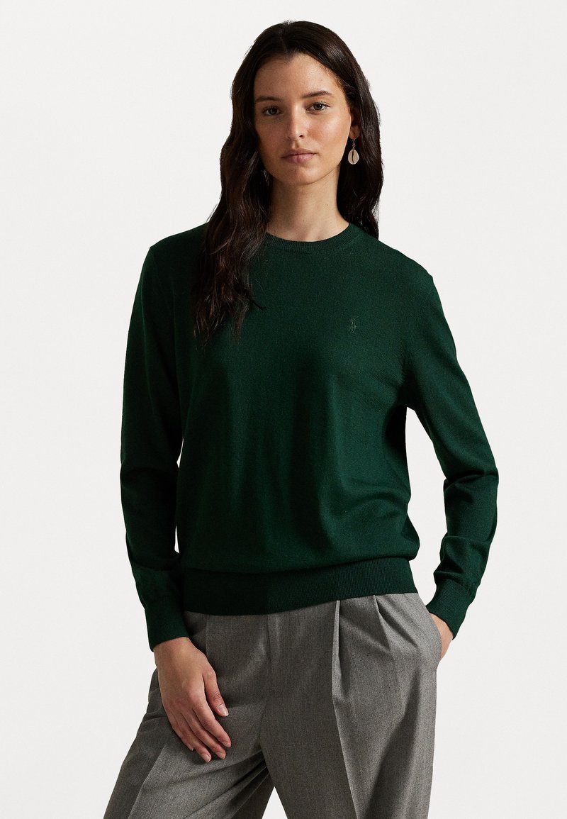 Green knit sweater with a crew neck, ribbed cuffs, and hem. Subtle logo detail on the chest. Paired with gray pleated trousers.