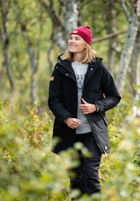 Veste imperméable noire avec capuche, associée à un pull motif gris clair, marchant à travers des feuillages, portant un bonnet bordeaux.