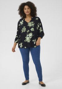 Zwarte bloemenblouse met groene rozen, met een v-hals, losse mouwen en een relaxte pasvorm, gecombineerd met blauwe skinny jeans en zwarte ballerina's.
