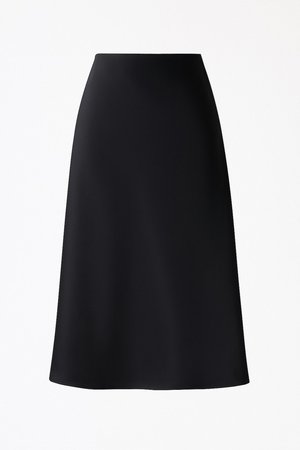 Melna A formas svārki, izgatavoti no gludas, mīkstas auduma. Raksturīga augsta jostasvieta un ceļgalu garuma apakša. Vienkāršs dizains bez redzamām rakstām vai akcentiem.