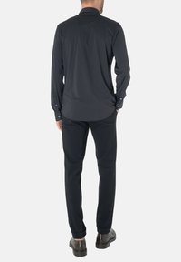 Maglietta nera a maniche lunghe con colletto all'italiana, tessuto morbido, polsini con due bottoni, abbinata a pantaloni neri e scarpe scure, vista dal retro.