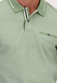 Lichtgroene polo met een gestructureerde brei, voorzien van een klassieke kraag, twee knopen en een zwart-witte streepaccent op de kraag. Kleine pocketdetail.