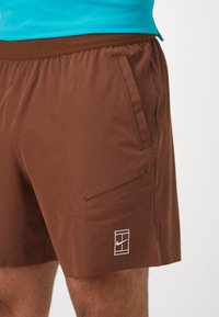 Brown athletic shorts with a flat waistband, side pocket, and white Nike logo. Smooth, lightweight fabric with a matte finish.