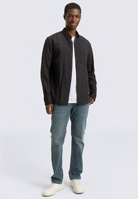 Schwarzes Hemd mit Knopfleiste und vertikalen Streifen, mit aufgesetzter Tasche, getragen über einem weißen T-Shirt, kombiniert mit hellblauen Jeans und weißen Sneakern.